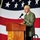 An elderly man in a gray suit and patterned tie is speaking at a wooden podium with a microphone. The podium bears the insignia and name of the USS Peleliu (LHA-5), a U.S. Navy amphibious assault ship. A large American flag is displayed prominently in the background.