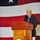 An elderly man is speaking at a wooden podium aboard the USS Peleliu (LHA-5), a U.S. Navy amphibious assault ship. The podium bears the ship's name, hull classification, and official seal with the motto 'Pax Per Potens' (Peace Through Power). A large American flag is displayed prominently in the background, and an audience is seated in the foreground.