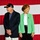 Two elderly individuals stand in front of a large American flag backdrop, suggesting a patriotic or political event. The man on the left wears a dark baseball cap with an American flag pin and a navy blazer, while the woman on the right wears a green cardigan over a white top with black pants. Both appear to be standing at attention or observing something off-camera.