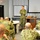 A military briefing or training session is taking place in a classroom-style setting. A single uniformed service member stands at the front of the room with arms crossed, addressing a group of seated personnel all wearing matching camouflage uniforms. The setting includes a whiteboard, projection screen, and podium, indicating a formal instructional environment.