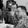 A black-and-white photograph showing three military personnel in digital camouflage uniforms seated at a table during what appears to be a briefing or meeting. The central figure is attentively listening with his hand raised to his chin in a contemplative pose. Computer monitors and office equipment are visible in the background.