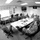 A military briefing is taking place in a conference room where uniformed personnel in camouflage uniforms are seated around a large table. One individual stands at the front, pointing to a whiteboard with a presentation displayed on it. The setting appears to be a formal military operations or planning meeting.