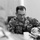 A U.S. Navy servicemember named McClelland, wearing Navy Working Uniform (NWU) digital camouflage, is seated at a table reviewing documents during what appears to be a briefing or meeting. He is wearing glasses and has rank insignia visible on his uniform. A coffee mug and papers are visible on the table in front of him.