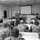 A military briefing or training session is taking place in a classroom-style auditorium. One uniformed service member stands at the front near a podium and projection screen, addressing a group of approximately 20-25 seated personnel all wearing matching camouflage uniforms. The setting appears to be a formal military instruction facility equipped with tiered seating, projection equipment, and multiple screens.
