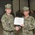 Two U.S. Navy personnel in NWU Type I (digital camouflage) uniforms are shaking hands while jointly holding a certificate reading 'Seabee Combat Warfare' during a formal military ceremony. The presenting officer, identified by name tape as 'McClelland,' appears to hold a senior rank indicated by a bird (eagle) insignia on his cap. A large formation of uniformed military personnel stands in the background inside what appears to be a hangar or large military facility.