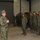A senior military officer in U.S. Navy Working Uniform (NWU Type III) addresses a formation of sailors inside what appears to be a hangar or warehouse facility. The officer stands facing the assembled personnel with hands clasped, appearing to deliver a briefing or speech. The formation consists of approximately 30-40 uniformed sailors standing at ease.