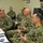 A group of U.S. Navy personnel in NWU (Navy Working Uniform) Type III camouflage are gathered in what appears to be a briefing or meeting room. Several senior personnel are seated around a table engaged in discussion, with additional personnel seated in the background. The setting appears to be an administrative or command office environment.