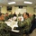 A group of approximately 10-11 military personnel in U.S. Navy digital camouflage uniforms (NWU Type I) are seated around a conference table in a briefing room. The setting appears to be a military installation, likely in Iraq based on the visible flag reading 'AL ASA &amp; IRAQ' on the back wall. A briefing or planning meeting is in progress with presentation materials visible.