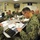 A group of U.S. Navy personnel in NWU (Navy Working Uniform) Type III camouflage are gathered in a briefing room at Al Asad, Iraq, as indicated by the flag/banner on the wall. They are seated around a conference table engaged in a formal briefing or planning session, with a projected presentation visible on the screen. A name placard reading 'McClelland' is visible on a portfolio on the table.