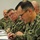 Several U.S. Navy personnel in NWU Type III (woodland digital camouflage) uniforms are seated at a table during what appears to be a briefing or meeting. The foreground figure is actively writing notes with a pen, while others in the background observe or take notes. The setting appears to be an indoor conference or training room.