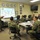 A military briefing is taking place in a conference room where a senior naval officer is presenting a slide titled 'FY 12/13 Operations' to a group of approximately eight personnel in Navy Working Uniform (NWU) camouflage. The presenter stands near a projection screen gesturing with both hands while the audience is seated around tables. The setting appears to be a formal operational planning or review meeting.