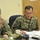 A U.S. Navy servicemember named McClelland is seated at a table during what appears to be a meeting or briefing. He is wearing the Navy Working Uniform (NWU) digital camouflage pattern and glasses, with documents in front of him. A second Navy servicemember is visible in the foreground with their back to the camera.