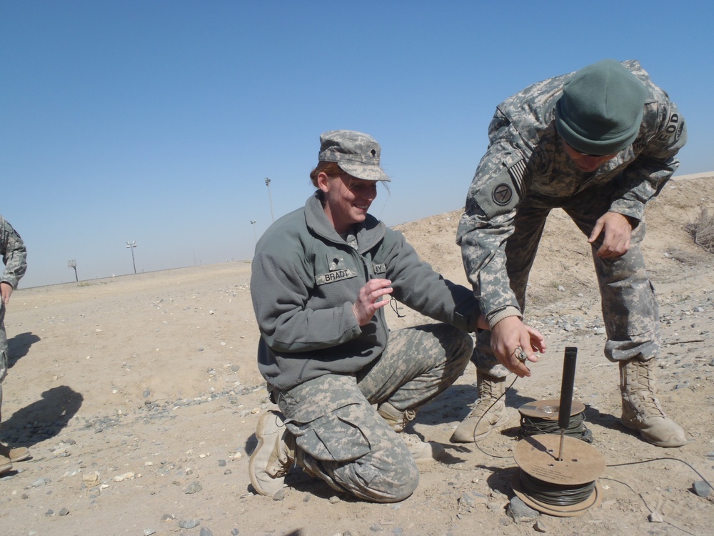 Two U.S. Army soldiers in ACU (Army Combat Uniform) are working with a spool of wire and a cylindrical device on arid, rocky terrain. The soldier on the left, identified by a name tape reading 'BRADT', is crouching and smiling, while the soldier on the right wearing a green beret is leaning over to handle the equipment. The setting appears to be a desert training or operational environment.