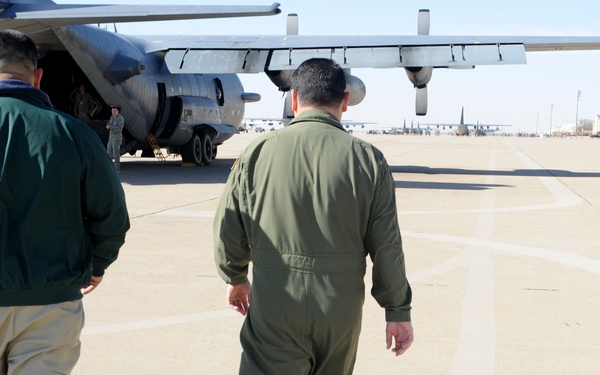 Two individuals are walking across a military airfield tarmac toward a large military transport aircraft. One person is wearing a green civilian jacket with khaki pants and has a prosthetic leg, while the other is dressed in an olive green military flight suit. Multiple military aircraft are visible in the background, indicating a large air base.