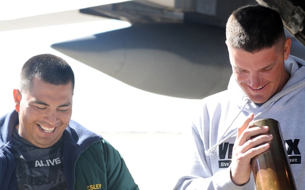 Two men are smiling and holding large brass artillery shell casings near what appears to be a military aircraft. The man on the left is wearing a green jacket with 'United States' insignia and the name 'LESLEY' visible, while the man on the right wears a gray hoodie with partial text 'VERTEX' visible. Both appear to be in good spirits, likely during a military base visit or tour.