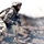 Big Island Military Police Training March 23-24 A U.S. Army soldier in full combat gear is moving through rough, disturbed terrain with smoke or dust visible in the foreground. The soldier is wearing ACU (Army Combat Uniform) digital camouflage and carrying a weapon while advancing through what appears to be a field exercise or combat environment. The image has a dramatic, high-contrast photographic style.