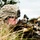 Big Island Military Police Training March 23-24 A soldier in U.S. Army ACU (Army Combat Uniform) digital camouflage is positioned low in tall grass, aiming a rifle with an underbarrel attachment. The soldier is wearing a combat helmet, tactical eyewear, and full combat gear. A military HMMWV (Humvee) vehicle is visible in the background, suggesting a field training exercise or operational deployment.