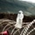 Big Island Military Police Training March 23-24 The image shows coils of razor wire (concertina wire) in sharp focus in the foreground, with a blurred human figure dressed in white standing behind the barrier in the background. The scene appears to be set in a rugged, hilly or mountainous landscape. The composition emphasizes the barrier as the primary subject, with the human figure suggesting themes of confinement, border control, or restricted movement.