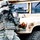 Big Island Military Police Training March 23-24 A soldier in U.S. Army ACU (Army Combat Uniform) digital camouflage is shown in a tactical stance, aiming a rifle while positioned near a tan-colored HMMWV (Humvee) military vehicle. The soldier is fully equipped with body armor, helmet, eye protection, and tactical gear. The image appears to be a training or demonstration scenario based on visible safety indicators on the weapon.