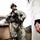 Big Island Military Police Training March 23-24 The image shows a U.S. Army soldier in full combat gear standing near a military vehicle, holding an assault rifle in a ready position. In the foreground, a partially visible individual wearing a white coat or robe with a decorative ring is visible. The composition suggests a military checkpoint or security interaction scenario.