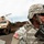 Big Island Military Police Training March 23-24 A U.S. Army soldier in full combat gear is positioned in the foreground, wearing ACU (Army Combat Uniform) digital camouflage, a tactical helmet, and ballistic sunglasses while carrying a rifle. In the background, a tan HMMWV (Humvee) military vehicle is visible along with at least one other soldier. The setting appears to be an outdoor field environment, likely a training exercise or operational deployment.