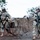 Big Island Military Police Training March 23-24 Two U.S. Army soldiers in ACU (Army Combat Uniform) pattern camouflage are deploying concertina/razor wire in a field environment. Both soldiers are armed and equipped with full combat gear, including helmets, body armor, and tactical vests. A military HMMWV (Humvee) vehicle is visible in the background.