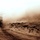 Big Island Military Police Training March 23-24 A military HMMWV (Humvee) is traveling along a dirt track in an arid, desert-like environment, kicking up a large dust cloud behind it. The sepia-toned photograph captures the vehicle from the rear as it moves away from the camera into a massive wall of dust. The sparse, dry vegetation and sandy terrain suggest an operational military environment, likely in a Middle Eastern or Central Asian region.