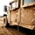 Big Island Military Police training A dusty military HMMWV (Humvee) is parked on sandy desert terrain, with a hand-drawn smiley face visible on its rear door panel. In the background, at least one soldier in full combat gear is visible in a kneeling or prone position. The image has a warm, desaturated tone consistent with arid desert environments.