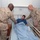 Two military personnel in desert camouflage uniforms are visiting a young man who is a patient in a hospital bed. The patient, wearing a blue Nike shirt, has a bandage on his nose and an IV line visible on his wrist. The uniformed individuals appear to be conducting a bedside visit, with one holding the patient's hand and the other placing a hand on his shoulder.