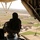 A military personnel is seated at the open rear ramp of a helicopter, looking down over a rural landscape with flat-roofed mud-brick structures and agricultural fields. An American flag is visible on the right side of the aircraft's interior. The image appears to be taken from a CH-47 Chinook or similar military transport helicopter during an aerial patrol or transit mission.