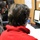 Two individuals are seated in what appears to be a radio or recording studio environment, both wearing headphones. The person in the foreground wears a red fleece hoodie and has shoulder-length dark hair, facing toward a computer monitor displaying what looks like audio software with a red recording indicator. A microphone is visible in the lower right corner of the frame.