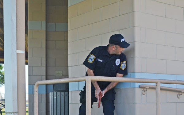 A uniformed police officer is standing near a railing at what appears to be a public park facility, holding what looks like a firearm at low ready. A person in civilian clothing (striped shirt, jeans, cowboy hat) is lying face-down on the ground. The setting appears to be a public restroom or pavilion structure at a park, with picnic tables visible in the background.