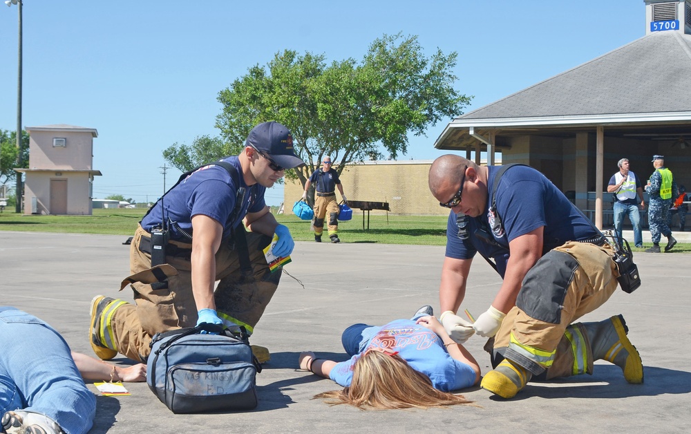 Two firefighters/EMTs in navy blue shirts and turnout gear are attending to a person lying on the ground in what appears to be a mass casualty drill or training exercise. A medical bag labeled 'NAS KING F.D.' is visible on the ground between them. Additional personnel, including individuals in military uniforms and safety vests, are visible in the background.