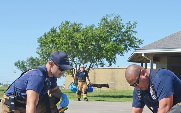 Two firefighters/EMTs in navy blue shirts and turnout gear are attending to a person lying on the ground in what appears to be a mass casualty drill or training exercise. A medical bag labeled 'NAS KING F.D.' is visible on the ground between them. Additional personnel, including individuals in military uniforms and safety vests, are visible in the background.