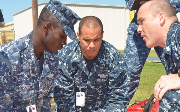 Three U.S. Navy personnel in NWU (Navy Working Uniform) digital camouflage are attending to a casualty lying on a red surface during what appears to be a medical training exercise or emergency drill. The sailors, identifiable by their name tapes and ID badges, are performing triage or first aid procedures on a person wearing a yellow shirt and jeans. A fire safety official in a high-visibility vest is visible in the background.