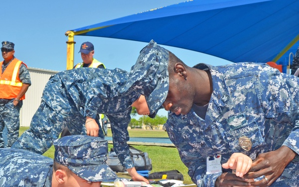 U.S. Navy personnel in blue digital camouflage uniforms are conducting a medical training exercise outdoors, attending to a simulated casualty lying on a stretcher. Several sailors are performing what appears to be first aid or triage procedures on the patient. Civilian safety observers in high-visibility vests are watching in the background.