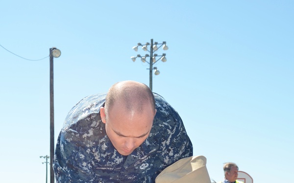 Two U.S. Navy personnel are providing medical treatment to a simulated casualty lying on a stretcher during what appears to be a mass casualty training exercise. One person is wearing Navy Working Uniform (NWU) digital camouflage and the other is in an olive green flight suit with a garrison cap and U.S. flag patch. Multiple other personnel in various uniforms are visible in the background participating in the same exercise.