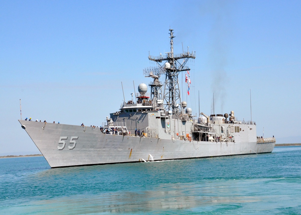 A gray naval warship bearing hull number '55' is photographed underway in calm, turquoise coastal waters under a clear blue sky. The vessel appears to be a Oliver Hazard Perry-class guided-missile frigate, identifiable by its characteristic mast configuration, radar domes, and hull design. Multiple crew members are visible on deck, and flags are flying from the mast.