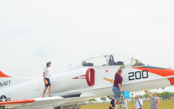 A group of approximately 10 people are washing and cleaning a U.S. Navy static display aircraft, identified as a T-2 Buckeye or similar trainer jet marked '200' with serial number 154338. The aircraft is painted in a white and orange training color scheme with 'NAVY' markings on the tail. The cleaning activity is taking place on a wet concrete pad, with volunteers using buckets, sponges, hoses, and ladders.