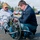 Two men are in a parking lot examining a hand-powered recumbent tricycle (handcycle). The man on the left is seated in the handcycle and wearing a U.S. Army cap and gray hoodie, while the man on the right is crouching down in a navy jacket and jeans, both focused on the bike's mechanics. The setting appears to be a commercial parking lot on an overcast day.