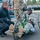 A U.S. Army soldier in ACU (Army Combat Uniform) is seated on a green hand cycle (recumbent handcycle) in a parking lot, wearing a black helmet and smiling. A man in Under Armour athletic wear is crouching beside him, appearing to converse or assist. A third person in military camouflage uniform is standing in the background.