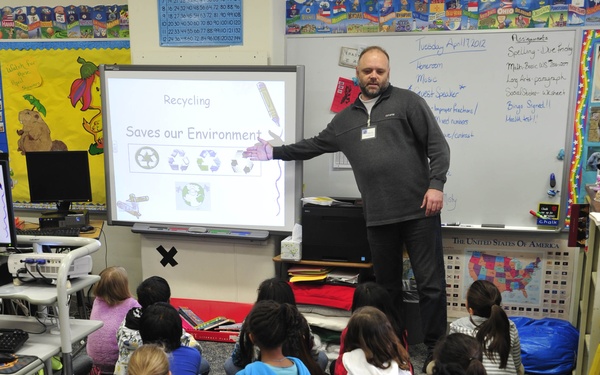 A male teacher or guest speaker is presenting a lesson on recycling to a group of elementary school students seated on the floor. The interactive whiteboard displays a slide titled 'Recycling Saves our Environment' featuring recycling symbols and environmental imagery. The classroom setting includes a whiteboard with the date 'Tuesday April 17, 2012' and various educational materials on the walls.