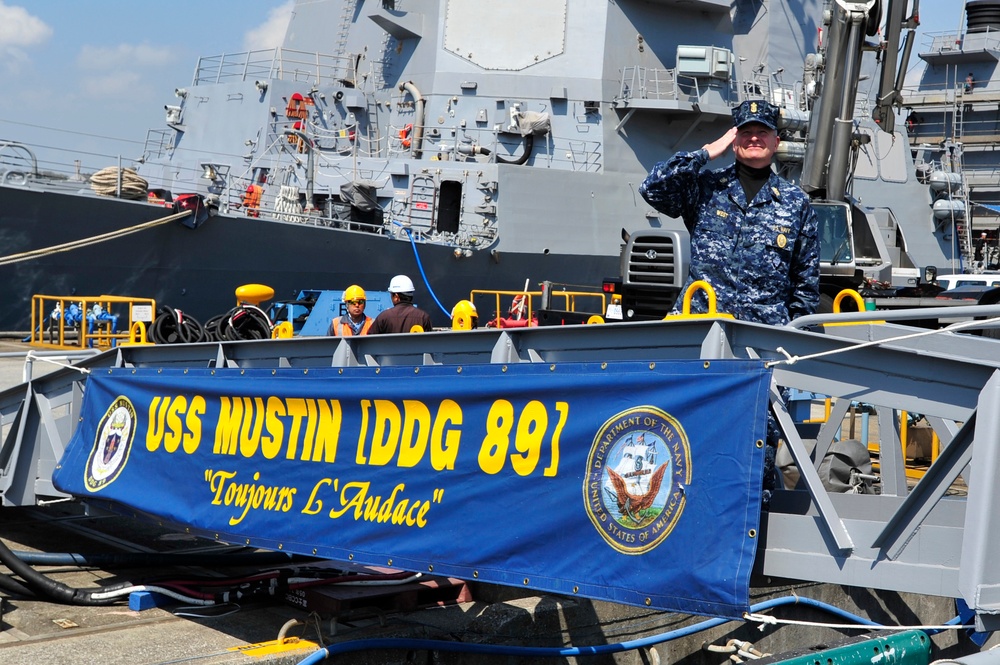 A U.S. Navy sailor in digital camouflage (NWU) uniform renders a salute aboard or near the gangway of USS Mustin (DDG 89), an Arleigh Burke-class guided-missile destroyer. A blue banner prominently displays the ship's name, hull number, and motto 'Toujours L'Audace' (Always Audacity) along with the U.S. Department of the Navy seal. The ship is moored at a pier with dockworkers visible in the background.