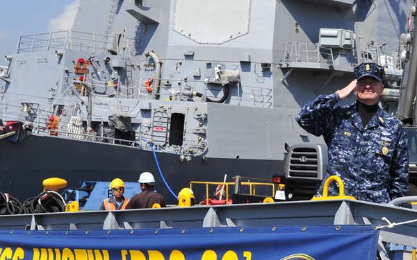 A U.S. Navy sailor in digital camouflage (NWU) uniform renders a salute aboard or near the gangway of USS Mustin (DDG 89), an Arleigh Burke-class guided-missile destroyer. A blue banner prominently displays the ship's name, hull number, and motto 'Toujours L'Audace' (Always Audacity) along with the U.S. Department of the Navy seal. The ship is moored at a pier with dockworkers visible in the background.