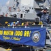 A U.S. Navy sailor in digital camouflage (NWU) uniform renders a salute aboard or near the gangway of USS Mustin (DDG 89), an Arleigh Burke-class guided-missile destroyer. A blue banner prominently displays the ship's name, hull number, and motto 'Toujours L'Audace' (Always Audacity) along with the U.S. Department of the Navy seal. The ship is moored at a pier with dockworkers visible in the background.