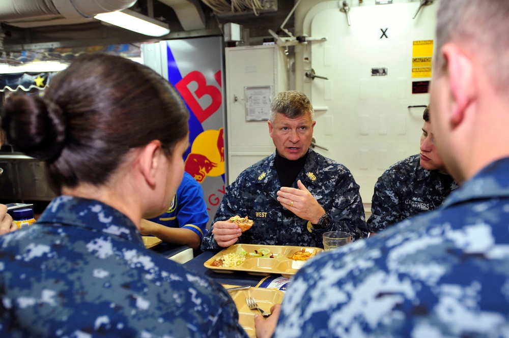 A group of U.S. Navy personnel in Navy Working Uniform (NWU) are seated together in what appears to be a ship's mess hall or galley. A senior officer, whose name tape reads 'WEST,' is speaking and gesturing while holding food, engaged in conversation with junior sailors. The setting includes standard military cafeteria trays with food and a Red Bull promotional display visible in the background.