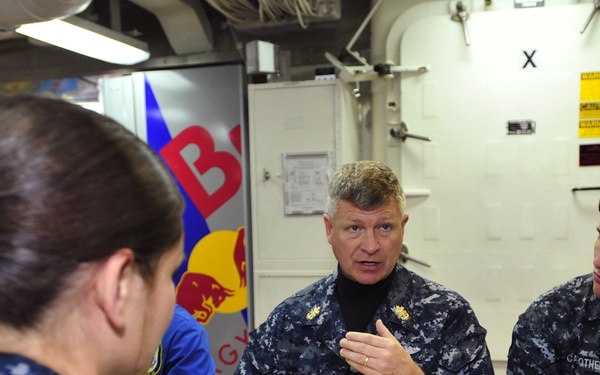 A group of U.S. Navy personnel in Navy Working Uniform (NWU) are seated together in what appears to be a ship's mess hall or galley. A senior officer, whose name tape reads 'WEST,' is speaking and gesturing while holding food, engaged in conversation with junior sailors. The setting includes standard military cafeteria trays with food and a Red Bull promotional display visible in the background.