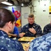 A group of U.S. Navy personnel in Navy Working Uniform (NWU) are seated together in what appears to be a ship's mess hall or galley. A senior officer, whose name tape reads 'WEST,' is speaking and gesturing while holding food, engaged in conversation with junior sailors. The setting includes standard military cafeteria trays with food and a Red Bull promotional display visible in the background.