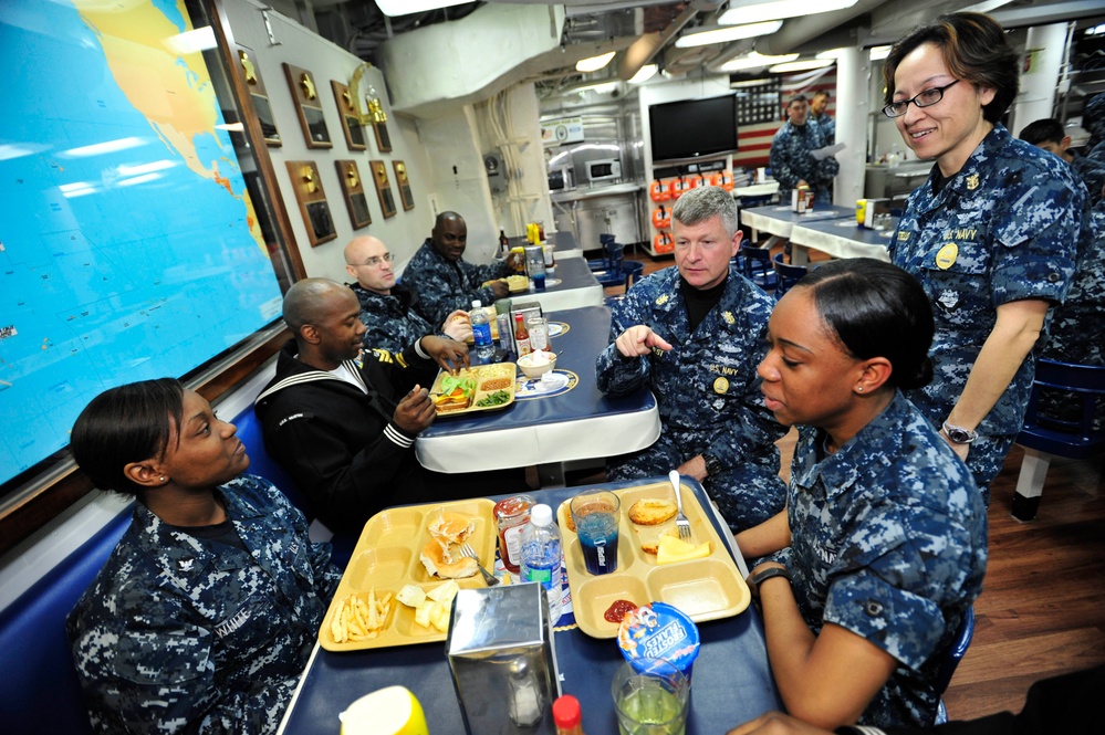 A group of U.S. Navy personnel in digital camouflage (NWU Type I) uniforms are gathered in what appears to be a ship's mess hall or wardroom. A senior officer is engaged in conversation with enlisted sailors seated at tables with food trays. The setting includes nautical charts, plaques on the walls, and an American flag in the background.