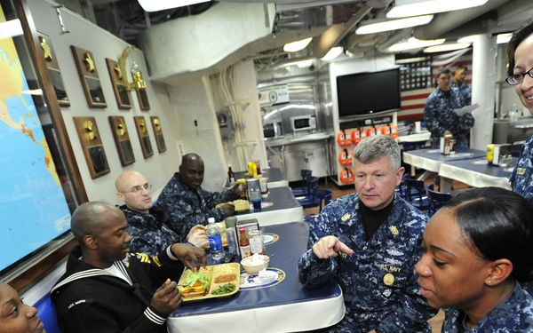 A group of U.S. Navy personnel in digital camouflage (NWU Type I) uniforms are gathered in what appears to be a ship's mess hall or wardroom. A senior officer is engaged in conversation with enlisted sailors seated at tables with food trays. The setting includes nautical charts, plaques on the walls, and an American flag in the background.